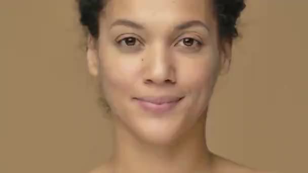 Portret de frumusețe al unei tinere afro-americane care se uită în oglindă, atinge fața și corectează părul. Modelul feminin negru pozând pe fundalul studioului maro. De aproape. Mișcare lentă gata, 4K la 59.94fps . — Videoclip de stoc
