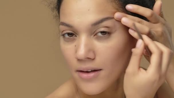 Portrait de beauté de jeune femme afro-américaine regardant dans le miroir et arrachant ses sourcils. Modèle féminin noir posant sur fond de studio brun. Ferme là. Prêt au ralenti, 4K à 59.94fps. — Video