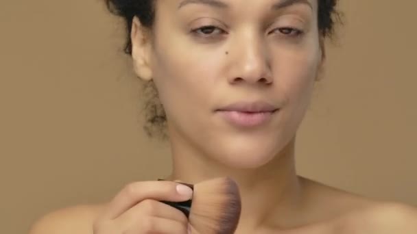 Retrato de belleza de una joven afroamericana que se aplica polvo en la cara con un cepillo de maquillaje. Modelo femenino negro posando sobre fondo de estudio marrón. De cerca. Cámara lenta lista, 4K a 59.94fps. — Vídeos de Stock