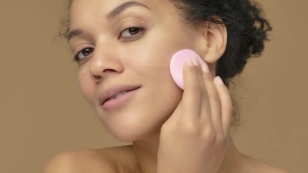 Krásný portrét mladé afroamerické ženy čistící tvář s růžovým diskem, odstranění make-upu. Černá žena pózuje na pozadí hnědého studia. Zavřít. Zpomalení připraveno, 4K při 59,94fps. — Stock video
