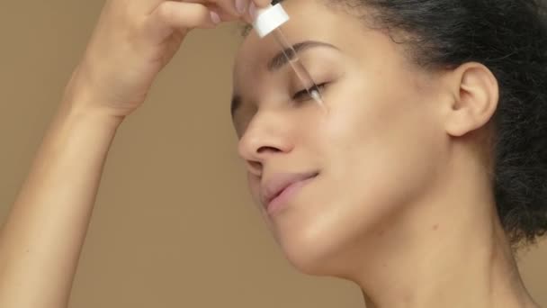 Retrato de belleza joven mujer afroamericana que aplica suero para el cuidado de la piel en la cara, frotando suavemente con los dedos. Mujer negra posando sobre fondo de estudio marrón. De cerca. Cámara lenta lista, 4K a 59.94fps. — Vídeo de stock