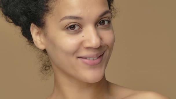 Beauty portret van jonge Afro-Amerikaanse vrouw smeert gezichtsverzorging crème op het puntje van haar neus en glimlacht gelukkig. Zwart vrouwelijk model op bruine studioachtergrond. Sluit maar af. Slow motion klaar, 4K bij 59,94fps — Stockvideo