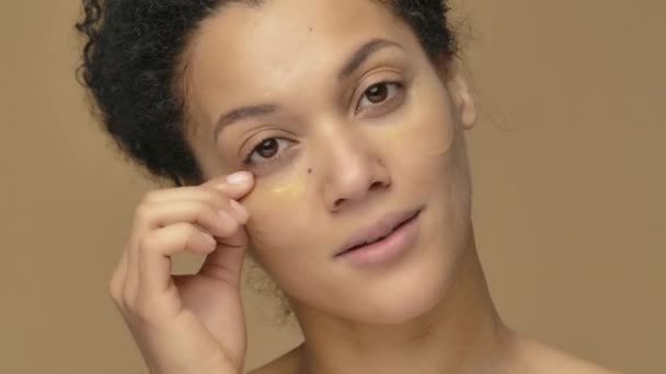 Schoonheidsportret van jonge Afro-Amerikaanse vrouw verwijdert ooggouden vlekken onder de ogen voor een frissere look. Zwart vrouwelijk model poserend op bruine studio achtergrond. Sluit maar af. Slow motion klaar, 4K bij 59,94fps. — Stockvideo