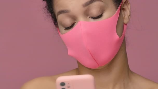 Retrato de beleza da jovem afro-americana em rosa máscara protetora pitta, mensagens de texto usando smartphone. Modelo nu feminino preto no fundo do estúdio rosa. Fecha. Movimento lento pronto, 4K a 59.94fps. — Vídeo de Stock
