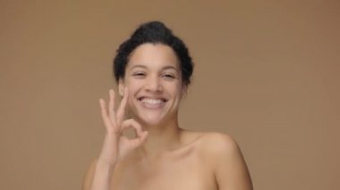 Genç Afro-Amerikan kadının güzel portresi gülümseyen, göz kırpan ve işaret gösteren. Kahverengi stüdyo arka planında poz veren siyah bir kadın model. Yavaş çekim hazır, 4K 59.94fps 'de..