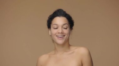 Genç Afrikalı Amerikalı kadının güzel portresi kameraya doğru dönüyor ve beyaz dişlerini göstererek gülümsüyor. Kahverengi stüdyo arka planında poz veren siyah bir kadın model. Yavaş çekim hazır, 4K 59.94fps 'de..