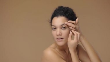 Genç Afro-Amerikan bir kadının aynaya bakıp kaşlarını yolmasının güzel portresi. Kahverengi stüdyo arka planında poz veren siyah bir kadın model. Yavaş çekim hazır, 4K 59.94fps 'de..