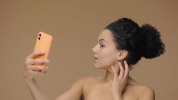 Retrato de belleza de una joven afroamericana tomando selfie usando un smartphone. Modelo femenino negro posando sobre fondo de estudio marrón. Cámara lenta lista, 4K a 59.94fps. — Vídeo de stock