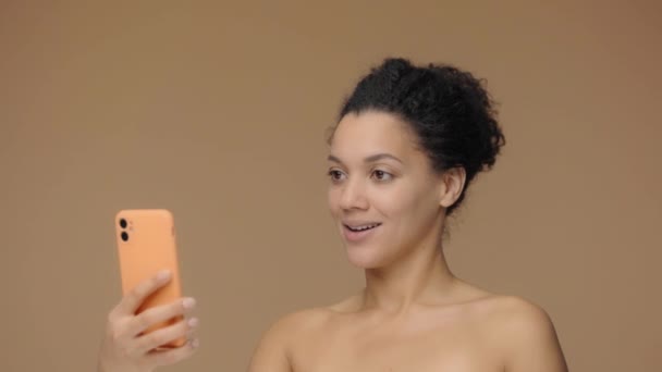 Krásný portrét mladé Afroameričanky mluví na videohovoru pomocí chytrého telefonu. Black female model pózuje na hnědém pozadí studia. Zpomalení připraveno, 4K při 59,94fps. — Stock video