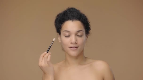Portrait de beauté de jeune femme afro-américaine regardant dans un miroir et se maquillant les yeux au pinceau. Modèle féminin noir posant sur fond de studio brun. Prêt au ralenti, 4K à 59.94fps. — Video