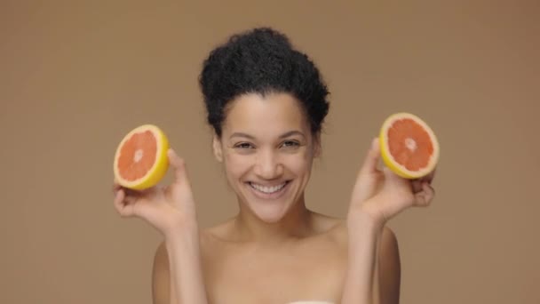 Schönheitsporträt einer jungen Afroamerikanerin, die Spaß hat und die Augen mit der Hälfte der Grapefruit bedeckt. Schwarze Hündin auf braunem Studiohintergrund. Zeitlupe bereit, 4K bei 59.94fps. — Stockvideo