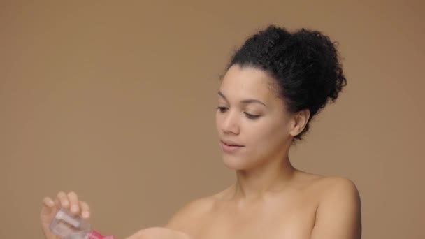 Krásný portrét mladé Afroameričanky, jak nalévá pleťovou vodu na podložku z bavlny a čistí obličej. Black female model pózuje na hnědém pozadí studia. Zpomalení připraveno, 4K při 59,94fps. — Stock video