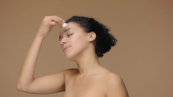 Ritratto di bellezza giovane donna afroamericana che applica siero per la cura della pelle sul viso, strofinando delicatamente con le dita. Donna nera in posa su sfondo marrone studio. Rallentatore pronto, 4K a 59.94fps. — Video Stock