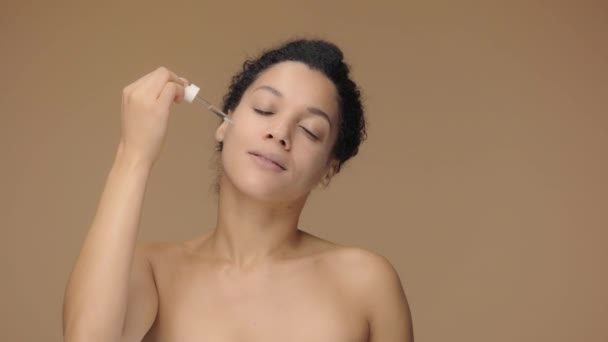 Retrato de belleza joven mujer afroamericana que aplica suero para el cuidado de la piel en la cara, frotando suavemente con los dedos. Mujer negra posando sobre fondo de estudio marrón. Cámara lenta lista, 4K a 59.94fps. — Vídeo de stock