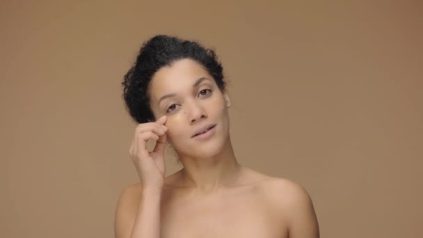 Skönhetsporträtt av ung afroamerikansk kvinna tar bort ögonguldfläckar under ögonen för fräschare look. Svart kvinnlig modell poserar på brun studio bakgrund. Slow motion klar, 4K vid 59.94fps. — Stockvideo