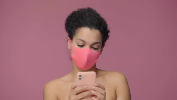 Schönheitsporträt einer jungen Afroamerikanerin in rosa Pitta-Schutzmaske, die per Smartphone SMS schreibt. Schwarzes weibliches Aktmodell auf rosa Studiohintergrund. Zeitlupe bereit, 4K bei 59.94fps. — Stockvideo