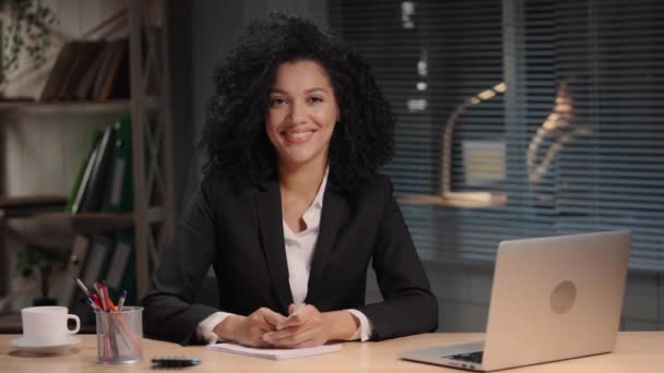 Portret van een Afro-Amerikaanse vrouw in een zakenpak die wegkijkt en glimlacht. Zakenvrouw poseren op het werk, zitten aan tafel in het interieur van het kantoor. Slow motion klaar, 4K bij 59,94fps. — Stockvideo