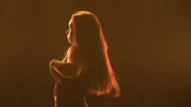 Siyah iç çamaşırlı, parlak ışıklarla karanlık bir stüdyoda dans eden zayıf bir kadının silueti. Dişi dansçı kalçasını sallar ve baştan çıkarıcı bir şekilde yarı çıplak vücudunu hareket ettirir. Kapat..