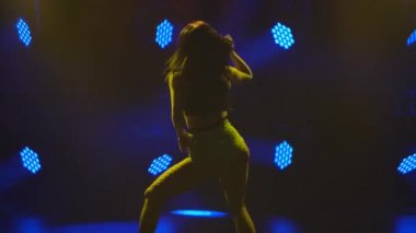 Kısa yeşil şortlu ve bluzlu seksi esmer kalçalı bir siluet. Şehvetli bir kadın, parlak ışıklarla dolu karanlık bir stüdyoda kalçalarını oynatıyor. Yavaş çekim.