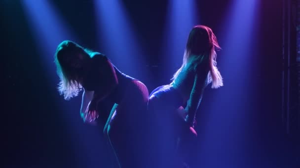 Two young hot women sexually move their bodies and twerk their ass. Silhouette of dancers in short shorts. Twerk duet on the background of blue lights. Slow motion. — Video