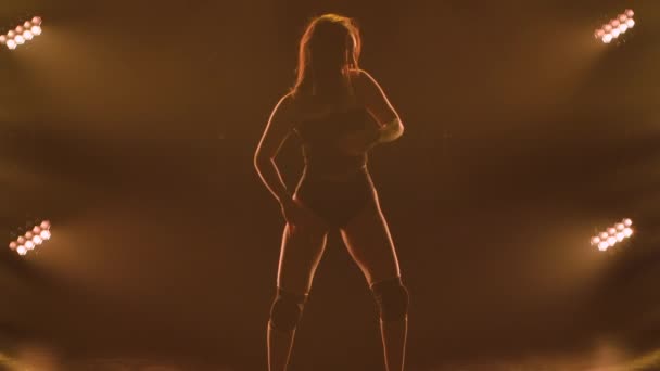 Silhouette of a sporty slender woman in black underwear dancing twerk in a dark studio with bright lights. The female dancer shakes her buttocks and seductively moves her half naked body. Slow motion. — Stock Video