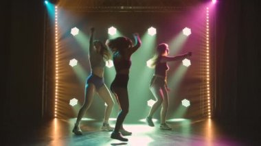 Group of three seductive dancers shaking their butts in short shorts. Silhouettes of young women twerking their asses moving their hips in a dark studio with bright lights. Slow motion.