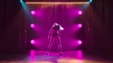 Sexy blonde shakes her ass seductively while dancing twerk in a dark studio with purple lights. Silhouette of a slender female body in black short shorts and a top. Slow motion.