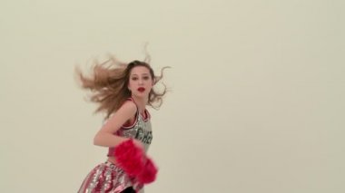 Beyaz stüdyo arka planında üniformalı bir ponpon kızın prova dansı. Amigo kız coşkulu bir dans eder, kollarını ve kalçalarını güçlü bir şekilde hareket ettirir, dikey bölünme yapar. Kapatın. Yavaş çekim.