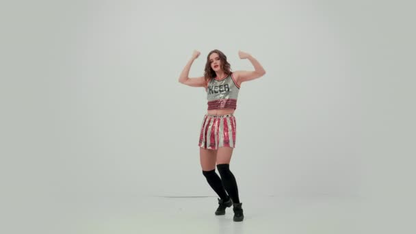 Solo performance of an attractive cheerleader in the studio on a white background. The girl energetically moves her arms and hips, demonstrating her plasticity, and sits on the twine. Slow motion. — Stock Video