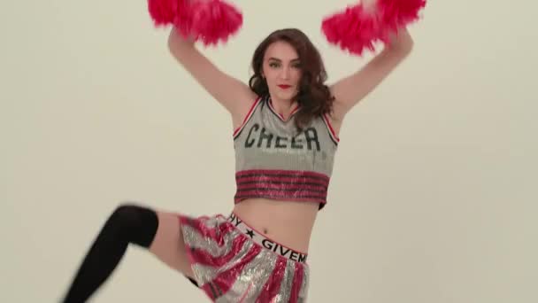 Young cheerleader with red pompoms in uniform is dancing on white background in studio. Beautiful cheerleader performs cheering dance. Pompons for cheerleading in female hands. Close up. Slow motion. — Stock Video