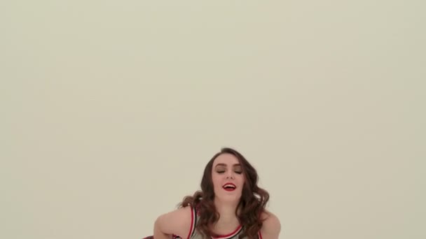 En flicka dansar en jublande dans med pom-poms i studion på en vit bakgrund. En hejaklacksledare räknar ut dansens element, rör på armarna, höfterna och hukar sig. Sakta i backarna. Närbild — Stockvideo