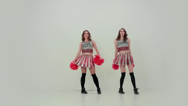 Dos animadoras uniformadas con pompones rojos bailan un baile edificante, mueven sus caderas. Las chicas bailan sincrónicamente en un fondo de estudio blanco en cámara lenta. Concepto de juegos de equipo, deporte profesional. — Vídeo de stock