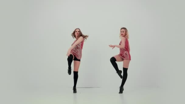 Twee cheerleaders in uniform dansen synchroon op een witte studio achtergrond in slow motion. Meisjes bewegen hun armen en heupen, demonstreren hun plasticiteit, zitten op het touw. Langzame beweging. — Stockvideo