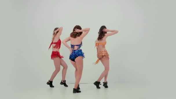 Trojice usměvavých žen tančí synchronně, pohybují rukama a boky a dělají svislé štěpení. Týmová práce roztleskávaček v lesklých kostýmech na bílém pozadí ve studiu. Zpomalený pohyb. — Stock video