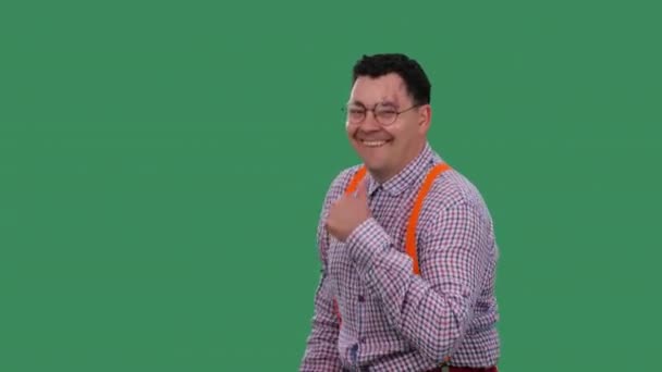 Задоволений чоловік танцює і показує великий палець руками, класом. Портрет чоловіка в окулярах, в плетеній сорочці з помаранчевими підтяжками в студії на зеленому екрані. Повільний рух. крупним планом . — стокове відео