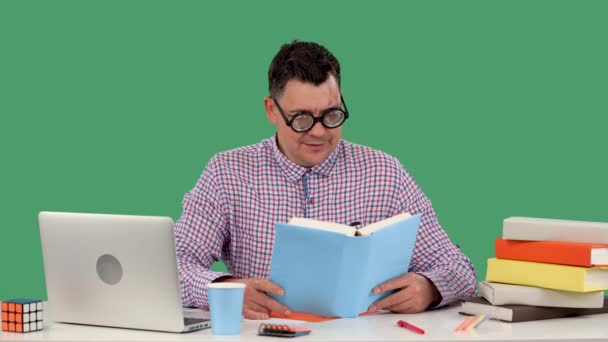 Um homem de óculos com lentes grossas senta-se em uma mesa na frente de um laptop e lê um livro, levanta seus óculos e diz. Retrato de um homem no estúdio numa tela verde. Movimento lento. Fechar. — Vídeo de Stock