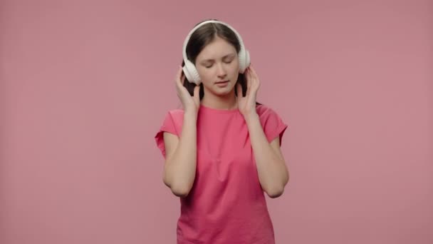 Potret model fashion menikmati musik dengan headphone besar berwarna putih nirkabel. Wanita berambut cokelat muda dengan rambut panjang berpose t-shirt pink di latar belakang studio merah muda. Tutup. Gerakan lambat siap 59.94fps — Stok Video