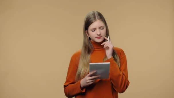 Retrato do modelo de moda pensando com concentração, escrevendo ideias no caderno. Menina com cabelos longos em camisola laranja posando no fundo do estúdio marrom. Fecha. Movimento lento pronto 59.94fps. — Vídeo de Stock