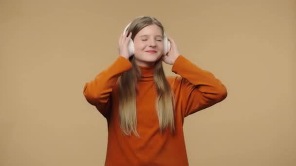 Porträtt av modemodell njuter av musik i stora trådlösa vita hörlurar. Ung snygg flicka med långt hår i orange tröja poserar på brun studio bakgrund. Närbild. Slow motion klar 59,94fps. — Stockvideo