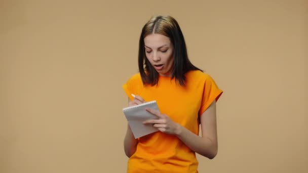Portrét módního modelu myšlení s koncentrací a psaní myšlenek a nápadů v notebooku. Mladá dívka ve žlutém tričku pózuje na hnědém pozadí studia. Zavřít. Zpomalení připraveno 59.94fps. — Stock video