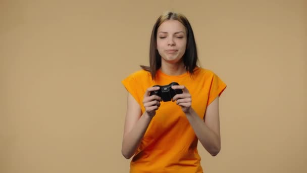 时尚模特儿运用无线控制器玩电子游戏的肖像,为胜利而欢欣.穿着黄色T恤的小女孩穿着棕色工作室背景的衣服。靠近点慢动作准备59.94fps. — 图库视频影像