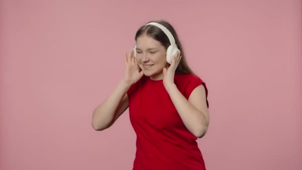 Porträtt av modemodell njuter av musik i stora trådlösa vita hörlurar. Ung flicka med långt hår i röd t-shirt poserar på rosa studio bakgrund. Närbild. Slow motion klar 59,94fps. — Stockvideo