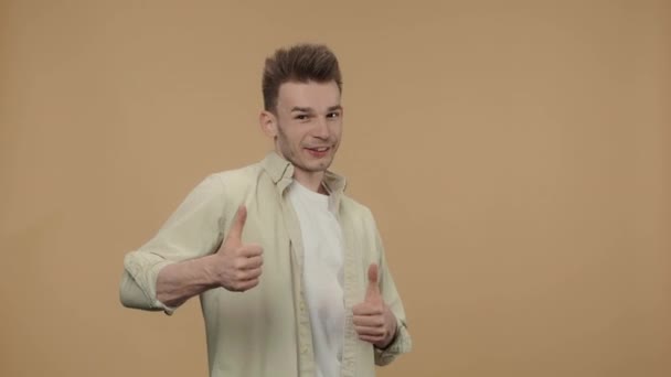Portrait of stylish guy showing thumbs up, gesture like and smiling. Male fashion model in a beige shirt posing on brown studio background. Close up. Slow motion ready 59.94fps. — Stock Video