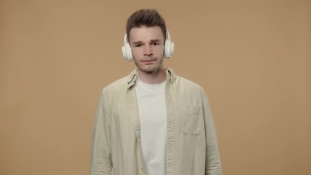 Portrait de type élégant appréciant la musique dans les grands écouteurs blancs sans fil. Modèle masculin dans une chemise beige posant sur fond de studio marron. Ferme là. Ralenti prêt 59.94fps. — Video