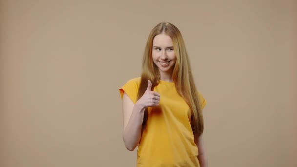 Porträtt av en modell med naturlig skönhet visar tummen upp, gest som och leende. Ung flicka med långt hår i gul t-shirt poser på brun studio bakgrund. Närbild. Slow motion klar 59,94fps. — Stockvideo