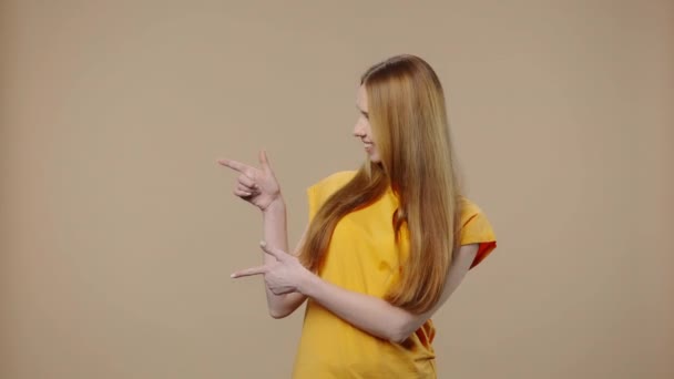 Porträtt av en skönhetsmodell pekar sida hand för något och visar tummen som. Ung flicka med långt hår i gul t-shirt poser på brun studio bakgrund. Närbild. Slow motion klar 59,94fps. — Stockvideo