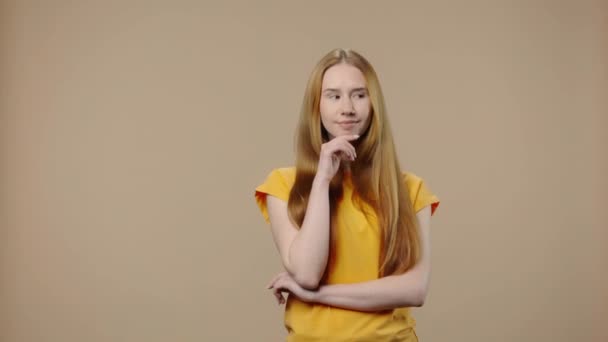 Retrato de un modelo con belleza natural pensando en algo con concentración. Chica joven con el pelo largo en camiseta amarilla posa sobre fondo marrón estudio. De cerca. Cámara lenta lista 59.94fps. — Vídeos de Stock