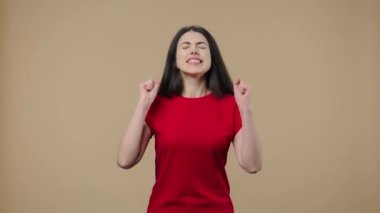 Doğal güzelliği olan bir modelin portresi kameraya heyecanla bakıyor ve zafer zaferini kutluyor. Genç esmer, kahverengi stüdyo arka planında poz veriyor. Kapatın. Yavaş çekim hazır 59.94fps.