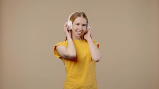 Porträtt av modell med naturlig skönhet njuter av musik i stora vita hörlurar. Ung flicka med långt hår i gul t-shirt poser på brun studio bakgrund. Närbild. Slow motion klar 59,94fps. — Stockvideo