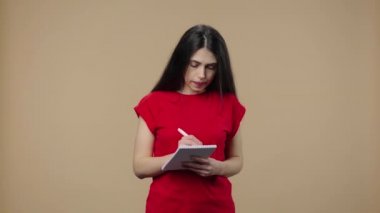 Güzellik modelinin portresi. Konsantrasyon ile düşünme. Not defterine fikirler yazmak. Uzun saçlı, kırmızı tişörtlü esmer kız kahverengi stüdyo arka planında poz veriyor. Kapatın. Yavaş çekim hazır 59.94fps